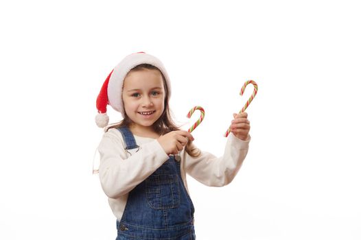 Adorable funny little girl in Santa hat and casual denim, smiling a beautiful toothy smile, looking at camera, pointing with Christmas lollipops at copy space for advertising text. Happy New Year Xmas