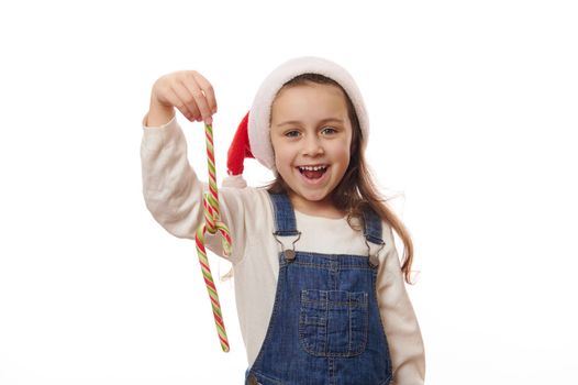 Caucasian smiling cheerful little girl wearing blue denim overalls and red Santa hat, playing with striped red and green candy canes lollipops on white background. Christmas New Year winter Holidays