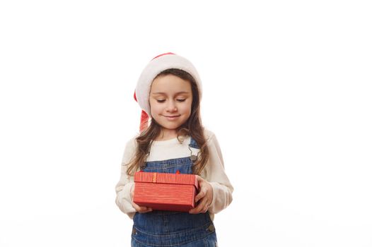 Isolated portrait on white background of a Caucasian cute little child girl in Santa hat, holding a red gift box with surprise for Christmas, expressing happiness and joy at upcoming winter holidays