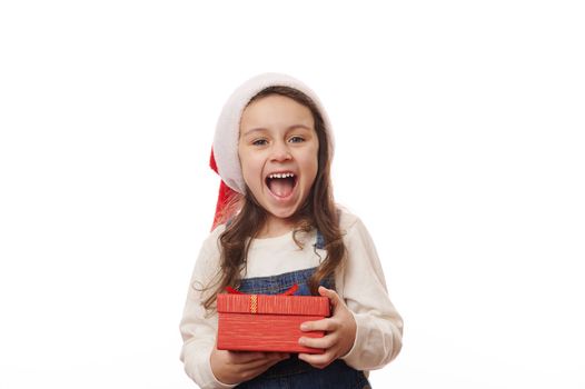 Happy overjoyed cheery child girl in Santa hat, rejoices looking at camera, holding a cute gift box for Xmas in her hands, isolated on white background with copy ad space. Merry Christmas. Boxing Day