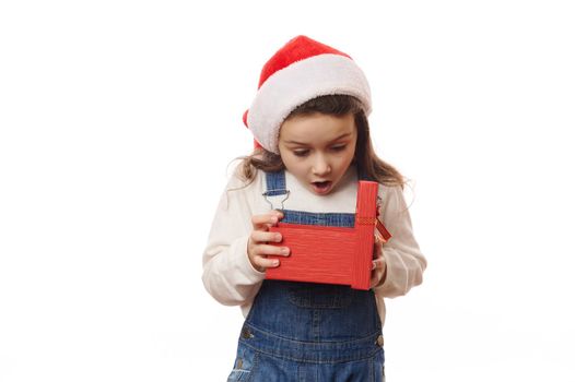 It's time to open Christmas New Year presents. Caucasian little girl in Santa hat, with excited emotions, unpacking a red gift box with cute surprise. Enjoy the magic atmosphere of winter holidays