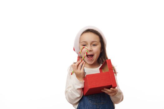 Caucasian 5-6 years old child, cheery baby girl in Santa hat, holding striped green red candy cane, rejoicing while unpacking Christmas presents, isolated over white background. Copy advertising space