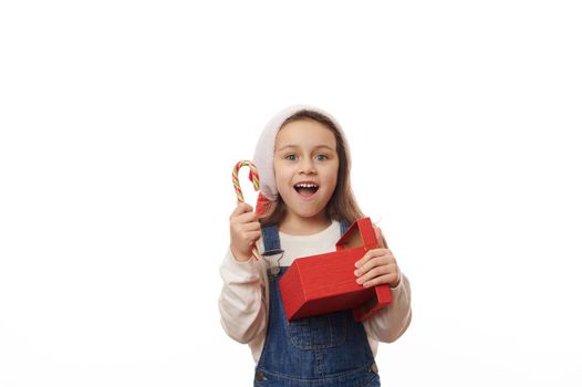Delightful baby girl in Santa hat, smiling a beautiul toothy smile looking at camera, posing with striped lollipop candy cane and red gift box, on white background. Merry Christmas and Happy New Year