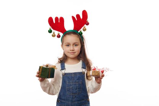 Cheerful Caucasian little girl in deer antler hoop, posing with happy Christmas presents over white background. Happy atmosphere of upcoming winter holidays. Boxing Day. It's time to open Xmas gifts
