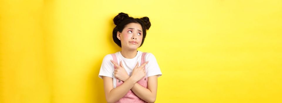 Indecisive asian teen girl troubled to make choice, pointing sideways and looking at logo confused, standing on yellow background.