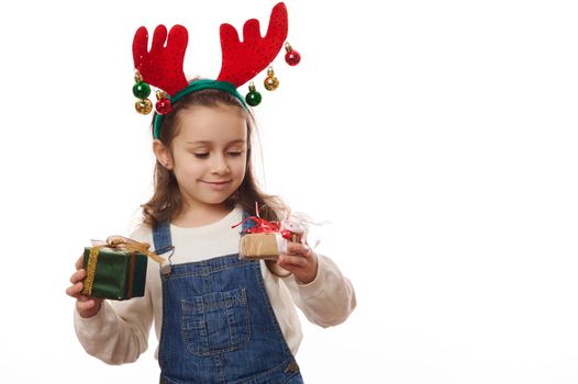 Happy Caucasian little girl wearing blue denim overalls and deer antler hoop, holding gift box with surprise for Christmas, cutely smiling over white background. Magical December mood. Copy ad space