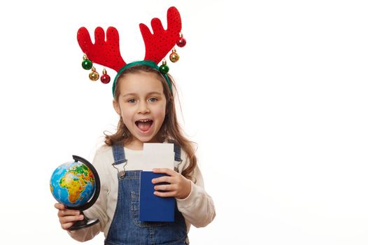 Adorable little girl wearing deer antler hoop, holding a globe and boarding pass, cheerfully smiling looking at camera, isolated over white background. Concept of travel abroad on winter holidays