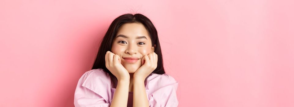 Close-up of excited asian girl listening with interest, smiling amused and looking at camera, standing against pink background.