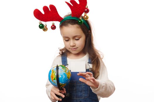 Charming Caucasian little girl in casual denim and deer antler hoop, posing with globe - the Earth planet over white background with copy ad space. Save Planet. Environmental conservation. Ecology