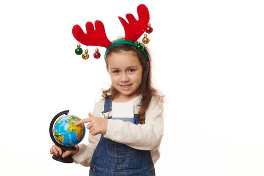 Happy child wearing deer antler hoop, pointing with forefinger at an Earth globe, making wish for saving planet against war damage, pollution. Environmental conservation concept on white background