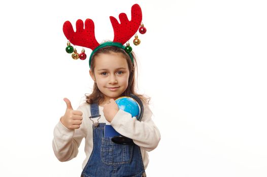 Caucasian child, lovely little girl in deer antler hoop, gently hugging the globe of Earth, showing thumb up looking at camera, isolated on white background. Kids loving planet. Copy advertising space
