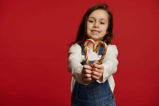Selective focus on green and red stripped lollipops - candy canes in the hands of a blurred child girl wearing white pullover and blue denim overrals, cutely smiling at camera, isolated red background