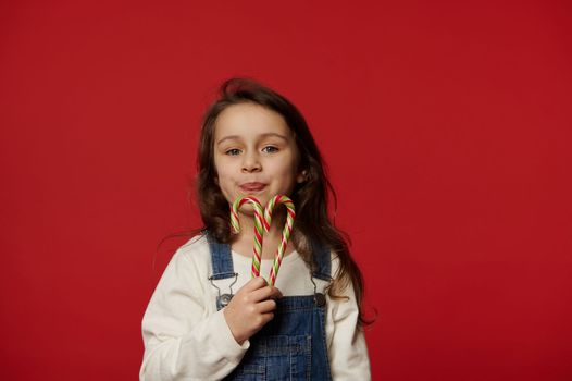 Beautiful little girl child holding sweet red striped green candy canes and cutely smiling looking at camera, isolated over red background with copy advertising space. Christmas and New Year concept