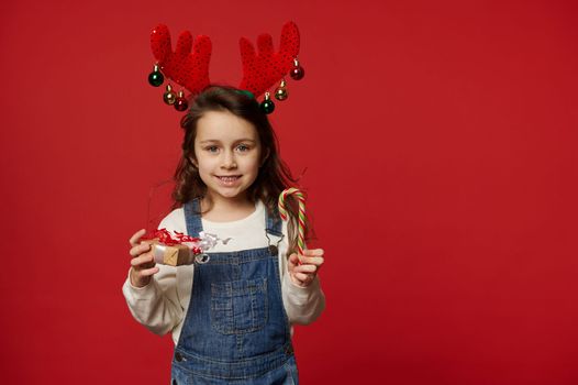 Adorable child, lovely little girl with long hair, wearing deer antler hoop and blue denim overalls, smiles looking at camera, posing with candy cane and Christmas present on red background Copy space