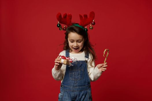 Adorable child little girl, wearing deer antler hoop, posing with stripped red and green lollipop - candy cane and Christmas present, rejoicing at upcoming winter holidays, isolated on red background