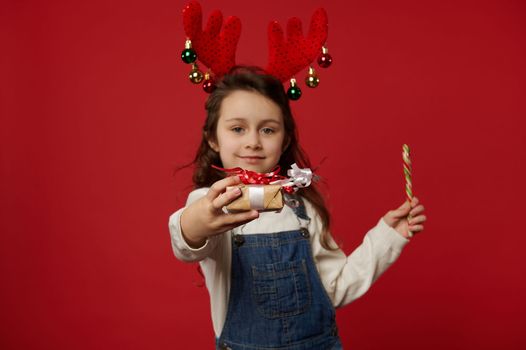 Focus on a cute gift, wrapped in craft wrapping paper and tied with red bow, in the hand of blurred little girl wearing deer antler hoop, holding out Christmas present at camera, over red background