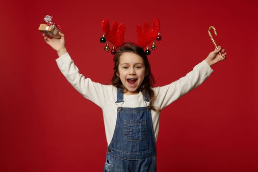 Overjoyed kid, lovely little girl in deer antler hoop, raising her arms up, holding Christmas gift and candy cane, isolated over red background with copy space, enjoying the upcoming winter holidays