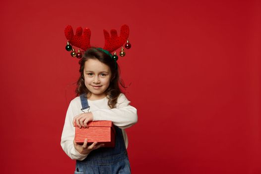 Adorable little girl wearing blue denim overalls, white pullover and deer antler hoop, unpacking a cute gift box with surprise present for Christmas, isolated on red background. Copy advertising space