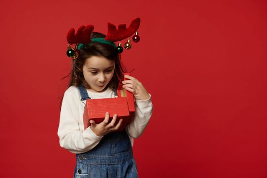 Surprised Caucasian baby girl wearing deer antler hoop, expressing happiness and excitement while unpacking red gift box with happy present for Christmas or New Year, over colored backdrop. Boxing Day