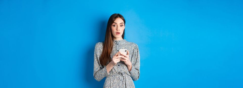 Woman look surprised after using mobile phone, standing on blue background.