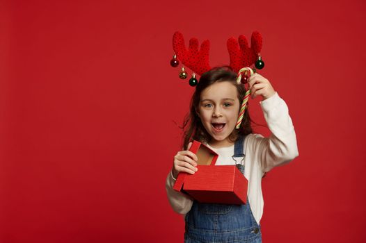 Adorable joyful and happy Caucasian little child, lovely baby girl wearing deer antler hoop, unpacking Christmas or New Year gifts and showing sweet candy cane, looking at camera, over red background