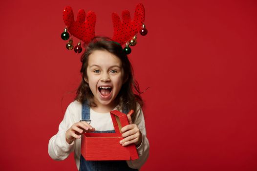 Overjoyed mischievous baby girl wearing denim overalls, white pullover and deer antler hoop, laughing looking at camera, expressing positive emotions, posing with Christmas present on red background