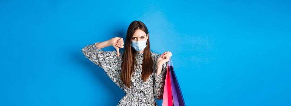 Covid-19, pandemic and lifestyle concept. Angry client in medical mask showing thumb down in dislike, frowning upset, holding shopping bags, blue background.