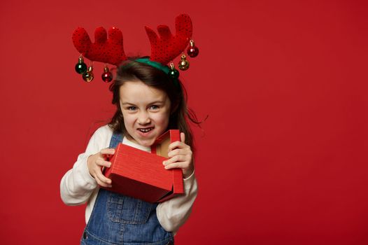 Mischievous funny little child girl in deer antler hoop, white pullover and blue denim overalls, smiling looking at camera, unpacking gift box with surprise for Christmas, on red background. Ad space