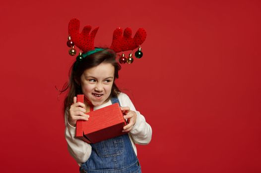 Mischievous cheery baby girl, wearing deer antler hoop, smiling looking aside at copy advertising space, unpacking Christmas or New Year gif box with presents, on red background. Happy winter holidays