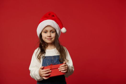 Lovely Caucasian little child girl in Santa hat, white pullover and blue denim overalls, smiling looking at camera, posing with cute gift box with surprise for Christmas, on red background. Ad space
