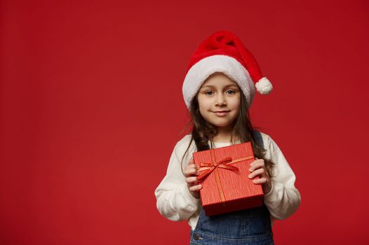 Caucasian 5-6 years old child, a lovely little girl in Santa Claus hat, white pullover and blue denim overalls, holding gift box with Christmas surprise, smiles at camera, isolated on red background