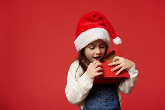 Delightful Caucasian 5 years old little child girl in Santa hat, expressing surprise and positive emotions while unpacking Christmas present over red background. Boxing Day. Happy New Year concept