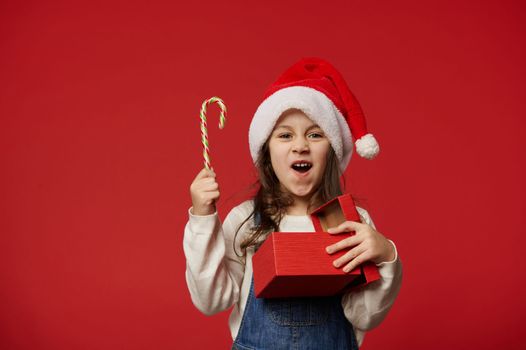 Surprised little girl wearing Santa hat, holding candy cane and red gift box with happy present for Christmas, expressing happiness and joy at upcoming winter holidays, red background. Merry Christmas