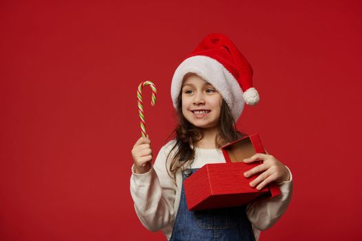 Caucasian happy 5 years old child, lovely little girl in Santa hat, smiling toothy smile looking at candy cane, posing with Christmas present on red background. Copy ad space. Winter holidays New Year