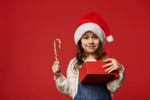 Beautiful little girl, happy child smiling looking at camera, holding sweet candy and gift box with happy present for Christmas or New Year, isolated on red background. Copy advertising space