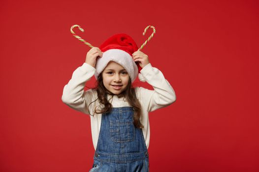 Joyful little girl with beautiful long hair, wearing white pullover, blue denim overalls and Santa Claus hat, holding striped lollipops on her head like horns, smiles looking at camera, red background