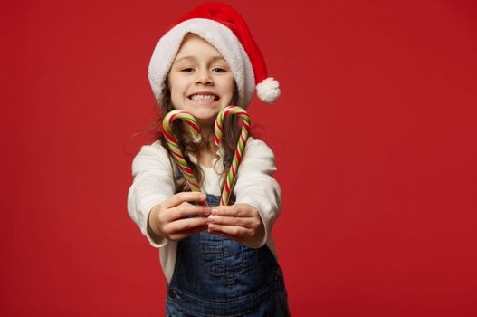 Details: hands of a blurred little child girl in Santa hat, smiling a beautiful cheerful toothy smile, holding out at camera red green stripped lollipops, sweet Xmas candy canes over red background