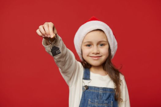 Focus on keychain in the hands of a delightful baby girl wearing Santa hat and blue denim overalls, holding out keys of new house or residence, cutely smiling looking at camera. Real estate concept