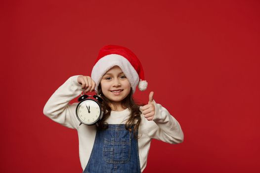 Caucasian cute little girl in Santa Claus hat, showing alarm clock with midnight on the dial and thumb up at camera, cutely smiling, expressing happiness, cheerfulness and positivity, red background