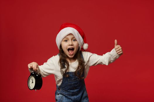 Overjoyed Caucasian 5 years old child girl with long hair, wearing Santa hat showing thumb up, posing with alarm clock showing midnight, looking at camera, red background. Happy New Year. Merry Xmas