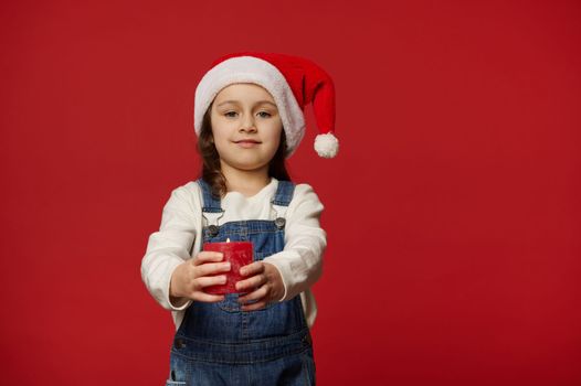 Caucasian lovely child, adorable blue-eyed little girl wearing Santa hat and blue denim overalls, cutely smiling looking at camera, holding out a lit Christmas candle, isolated on red background