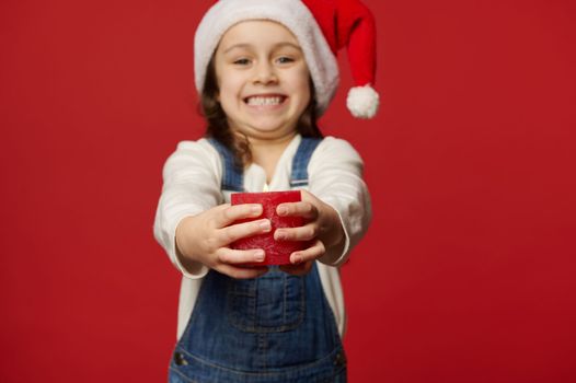 Selective focus on lit Christmas candle in the hands of blurred cheery child girl in Santa hat, smiling a cheerful toothy smile looking at camera, isolated over red background. New Year's celebration