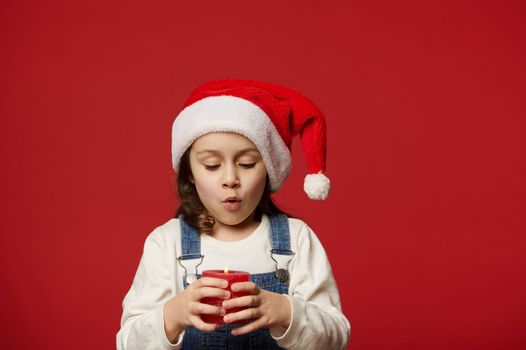 Adorable Caucasian little child girl wearing blue denim overalls, white pullover and Santa hat, blowing out a candle, making cherished wish for Christmas on red background. Happy New Year. Copy space