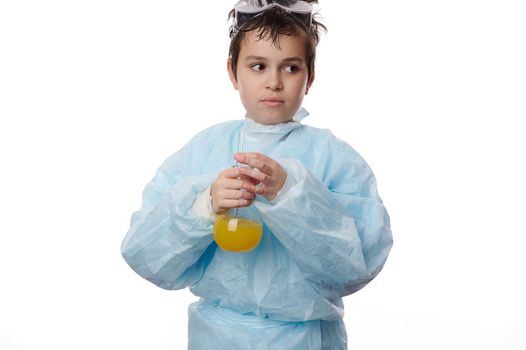 Multi-ethnic thoughtful smart teenage schoolboy in lab coat, holding a glass flask with yellow chemical liquid substance, doing experiments, isolated over white background. Chemistry Science concept
