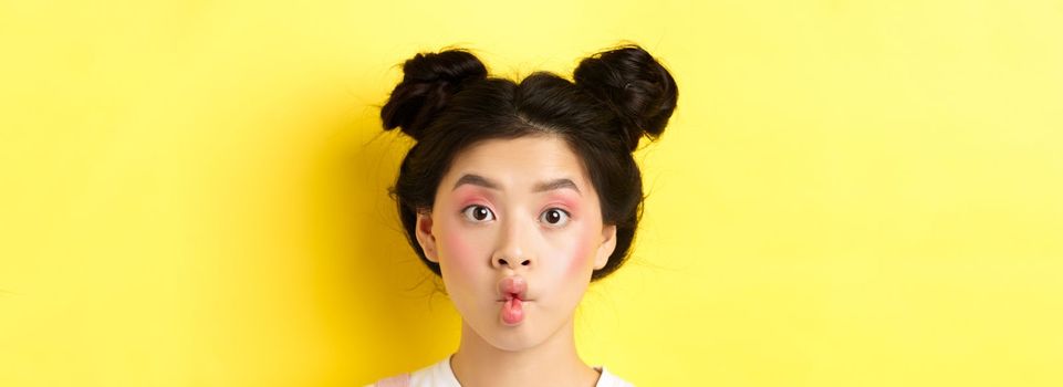 Close-up of teen korean girl pucker lips and looking funny at camera, standing with glamour makeup and stylish hairstyle, yellow background.