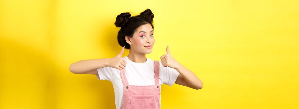 Beautiful asian girl with bright pink makeup, showing thumb up in approval and smiling satisfied, recommending promo, yellow background.