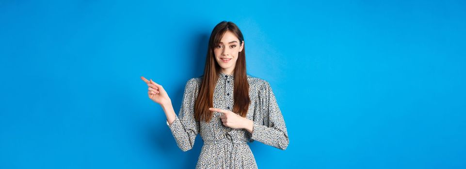 Young attractive woman in dress, with long natural hair, pointing fingers right at logo, smiling and inviting check out promotion, standing on blue background.