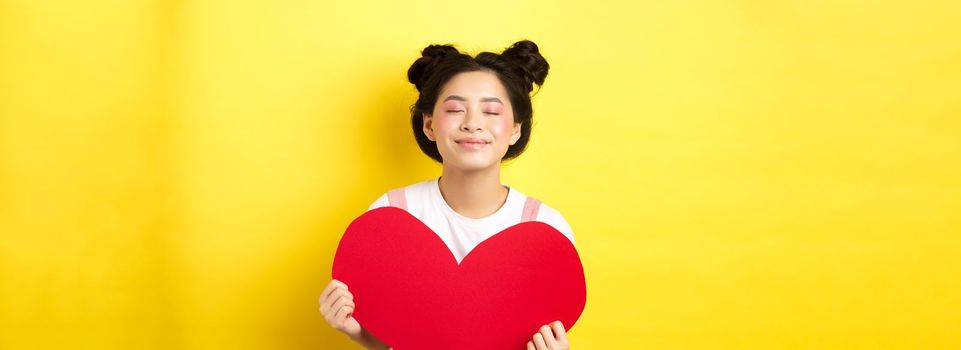 Cute teenage asian girl dreaming of true love, close eyes and showing big red heart cutout, waiting for soulmate on Valentines day, standing with romantic makeup on yellow background.