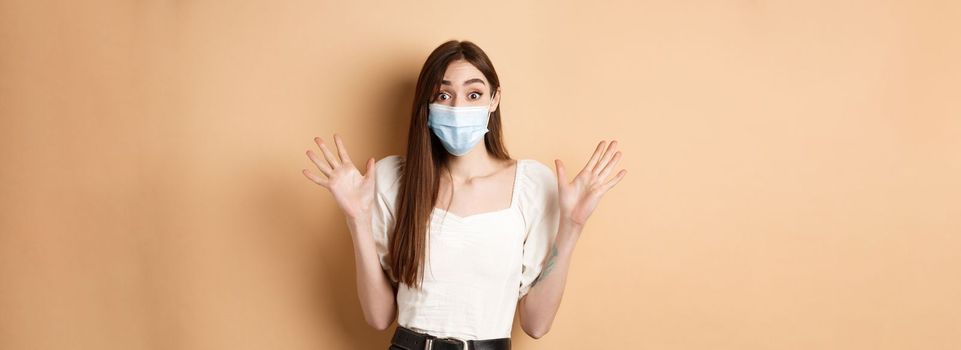 Covid-19 and lifestyle concept. Surprised woman in medical mask scream excited, raising hands up and shouting big news, standing on beige background.