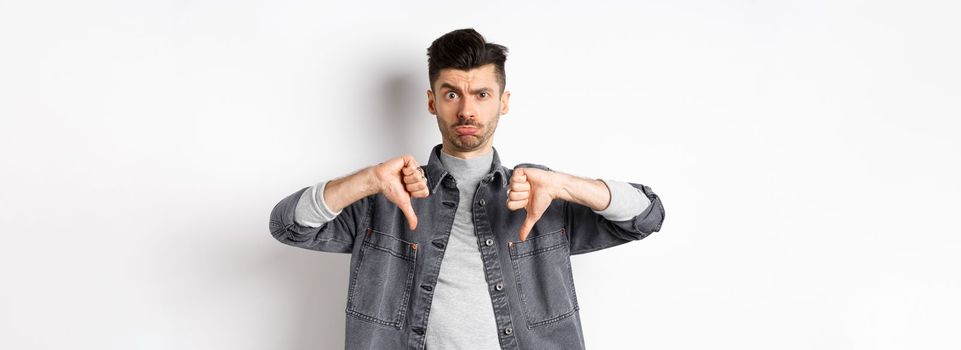 Disappointed funny man frowning and looking upset, showing thumbs down displeased, standing on white background.